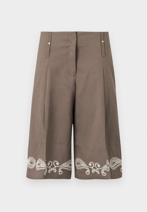 Nő, aki barna ujjatlan, gombos mellényt és hozzáillő, fehér hímzéssel díszített térd alatti rövidnadrágot visel, világos bézs színű sportcipővel párosítva.