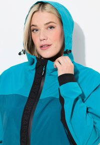 Veste imperméable teal avec capuche, présentant des accents noirs et une fermeture éclair. Le matériau a une texture lisse et un design sportif.