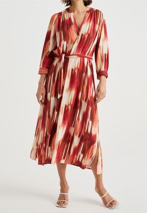 Frau trägt ein langärmeliges Midikleid mit Gürtel, bedruckt mit einem abstrakten Muster in Rot, Weiß und Beige und einem seitlichen Schlitz, kombiniert mit weißen Sandalen.