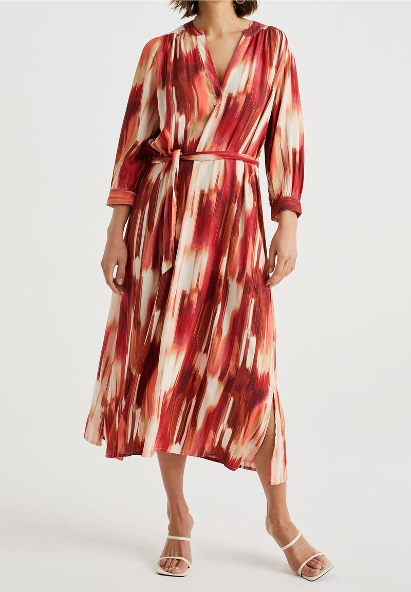 Femme portant une robe midi à manches longues ceinturée, avec un motif abstrait de coups de pinceau rouges, blancs et beiges, fendue sur le côté, associée à des sandales blanches.
