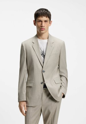 Jeune homme portant un costume beige clair à deux boutons, un T-shirt blanc graphique en dessous, debout avec une main dans la poche sur fond blanc.