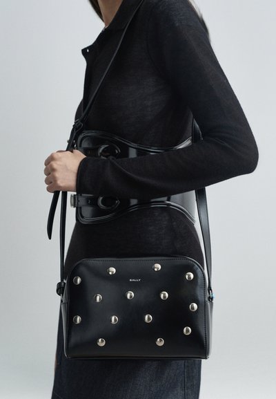 Černá kožená kabelka s kovovými nýty, kompaktní tvar, nastavitelný popruh a detaily loga. Nošena s černým přiléhavým topem a lesklým opaskem.
