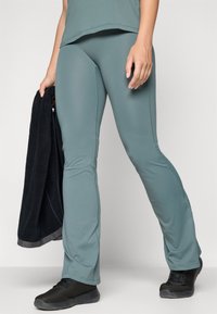 Teal treningstights i elastisk materiale med vide ben og sidesømmer. Kombinert med et svart håndkle og svarte joggesko.