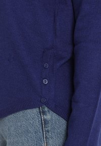 Granatowy dzianinowy kardigan z okrągłym dekoltem, bocznymi guzikami, ściągaczami przy rękawach i lekko asymetrycznym dołem. Noszony na denimie.