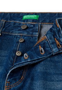 Dżinsy w kolorze niebieskim, z metalowymi guzikami i pomarańczowymi szwami; metka głosi "United Colors of Benetton." Dwa haftowane otwory na guziki.