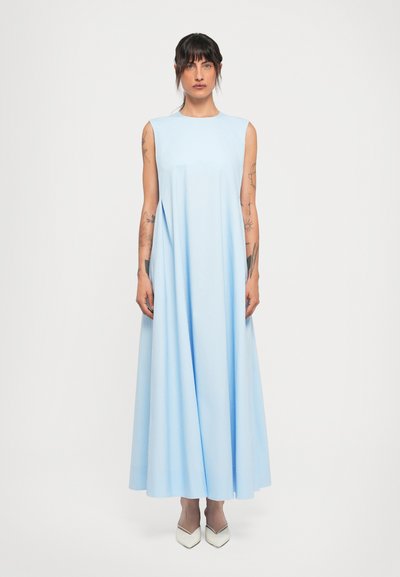 Vrouw met donker haar en tatoeages, gekleed in een mouwloze lichtblauwe maxi-jurk en witte puntige schoenen, staande tegen een witte achtergrond.