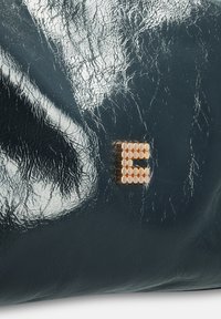 Donkerzeegroene, glanzende neplederen tas met een gestructureerd oppervlak en een gouden metalen letter "E" bestaande uit ronde studs.