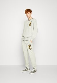 Helles graues Zweiteiler-Outfit bestehend aus einem Langarmoberteil mit V-Ausschnitt und gesticktem Logo, kombiniert mit passenden Jogginghosen und grauen Sneakern.