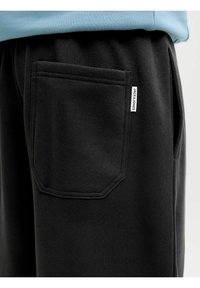 Pantalones de chándal negros con un gran bolsillo trasero cuadrado y costuras geométricas, hechos de una tela suave. Se puede ver una pequeña etiqueta en el costado.