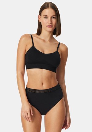 Donna che indossa un reggiseno senza cuciture nero con spalline sottili e slip a vita alta con fascia in rete, in piedi davanti a uno sfondo neutro.