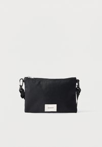 Čierna textilná kabelka cez rameno so zipsovým uzáverom, nastaviteľným popruhom a bielou značkovou etikietou na prednej strane. Jednoduchý obdĺžnikový dizajn.