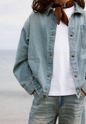 Osoba nosiaca svetlomodrú džínovú bundu a rifle, biele tričko a hnedý šál okolo krku, ruky v kapsách, blízko vodnej plochy.