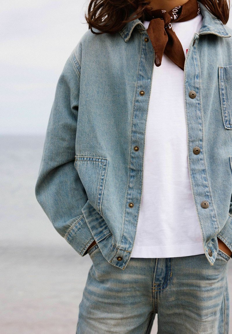 Osoba nosiaca svetlomodrú džínovú bundu a rifle, biele tričko a hnedý šál okolo krku, ruky v kapsách, blízko vodnej plochy.