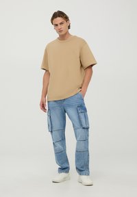 T-shirt oversize beige abbinato a jeans cargo blu con più tasche, completato da sneakers bianche. Design semplice e casual.