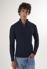 Pull en mailles torsadées bleu marine avec col montant et demi-fermeture éclair, doté de poignets et d'ourlet côtelés, porté par-dessus un jean bleu clair.