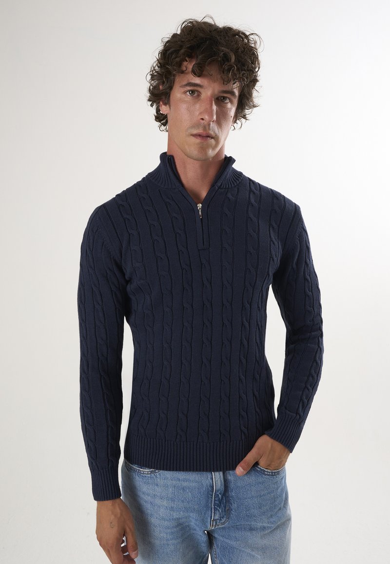 Pull en mailles torsadées bleu marine avec col montant et demi-fermeture éclair, doté de poignets et d'ourlet côtelés, porté par-dessus un jean bleu clair.