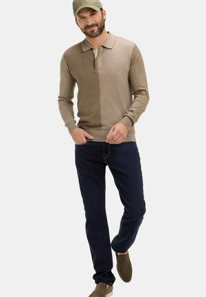 Homme portant un polo à manches longues bicolore beige, un jean bleu foncé, des chaussures à enfiler olive et une casquette olive, debout les mains près des poches.