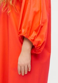 Χέρι σε φωτεινό πορτοκαλί φόρεμα με φουσκωτά μανίκια και ελαστικό μανσέτα, ανοιχτόχρωμος τόνος δέρματος και σγουρά ξανθά μαλλιά ορατά στην επάνω αριστερή πλευρά.
