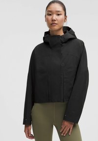 Kvinde iført en sort hættejakke og olivengrønne leggings, der står mod en ensfarvet lys baggrund, vender fremad med et neutralt udtryk.