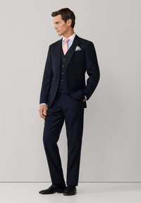 Mężczyzna ubrany w granatowy garnitur trzyczęściowy, jasnoniebieską koszulę, różowy krawat oraz poszetkę w grochy, stoi z jedną ręką w kieszeni.