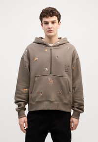 Sudadera con cremallera color marrón hecha de algodón, que presenta varios motivos de animales bordados en naranja, blanco y marrón. Corte holgado con un bolsillo tipo canguro.