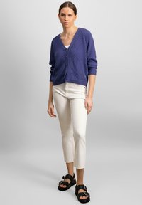 Fialový pletený sveter s výstrihom do V a zapínaním na gombíky, skombinovaný s krémovými skrátenými džínsami a čiernymi sandálmi so strapcami.