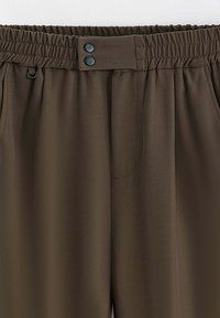 Pantalon en tissu marron léger avec taille élastique, deux poches latérales et deux boutons décoratifs à la taille. Texture lisse.