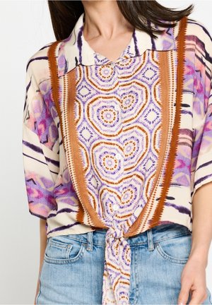 Femme portant une chemise à manches courtes à boutons avec des motifs géométriques violets, orange et roses nouée à la taille, associée à un jean bleu clair.