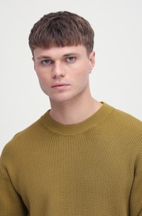 Musztardowo-żółty sweter o fakturowanej strukturze z szerokim dekoltem; na modelu z krótkimi włosami i kolczykiem, na jasnym tle.