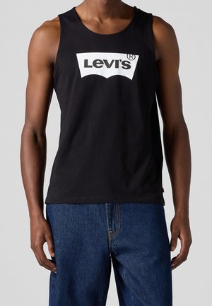 Mand iført sort Levi's ærmeløs tanktop og blå jeans foran en ensfarvet grå baggrund.