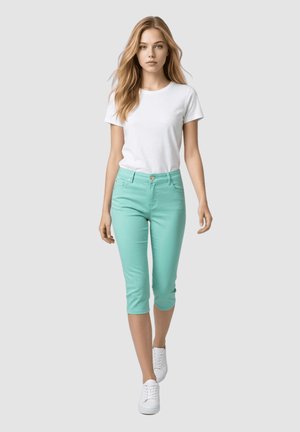 Joven mujer caminando hacia adelante con camiseta blanca, pantalones capri verde menta y zapatillas blancas sobre un fondo liso gris claro.