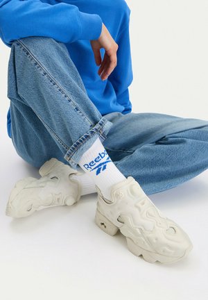 Persona con jeans azules y sudadera azul sentada con las piernas cruzadas, mostrando calcetines blancos Reebok y zapatillas gruesas beige.