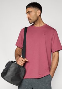 Ανδρικό κοντομάνικο μπλουζάκι σε μοβ απόχρωση με στρογγυλή λαιμόκοψη και λογότυπο της Nike. Φοράει μια μαύρη, δομημένη τσάντα με ρυθμιζόμενο ιμάντα.