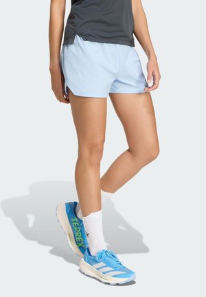 Persona che indossa pantaloncini sportivi azzurri chiaro, calzini bianchi e scarpe da corsa Adidas Terrex blu, in piedi con una gamba leggermente piegata.