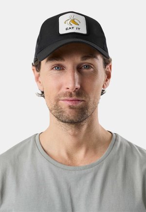 Mann mit leichtem Bart, der eine schwarze Mütze mit einer Grafik einer geschälten Banane und den Worten „EAT IT“ trägt, sowie ein hellgraues Rundhalsshirt.