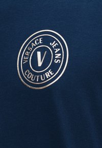 Μεταλλικό ασημί λογότυπο Versace Jeans Couture με ένα έντονο "V" μέσα σε δύο ομόκεντρους κύκλους σε σκούρο μπλε ύφασμα.