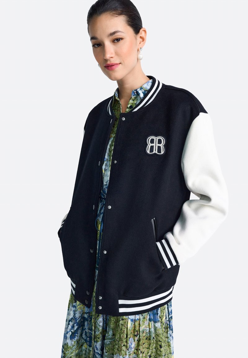 Fekete-fehér egyetemi stílusú dzseki gyapjú testtel, bordázott mandzsettákkal és hímzett logóval. Virágmintás ruha fölött viselve. Sima tapintású.