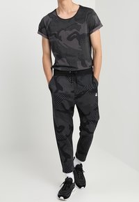 Černé tričko s krátkým rukávem v maskáčovém vzoru, doplněné černými joggerkami s šňůrkou v pase a bočními pruhy, nošené s černými teniskami.