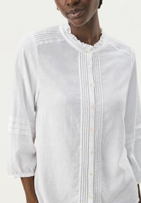 Witte linnen blouse met een rimpel halslijn, knopen aan de voorkant en verticale plooien. Driekwart mouwen met horizontale strepen bij de boorden.