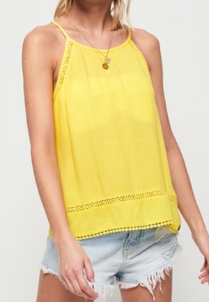 Żółta bezrękawnikowa bluzka z lekkiego materiału, z wykończeniem z koronki na dole, zestawiona z postrzępionymi szortami jeansowymi.