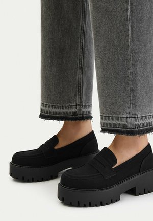 Mocasines de plataforma gruesos negros usados con vaqueros grises recortados con bajos deshilachados sobre un fondo claro.