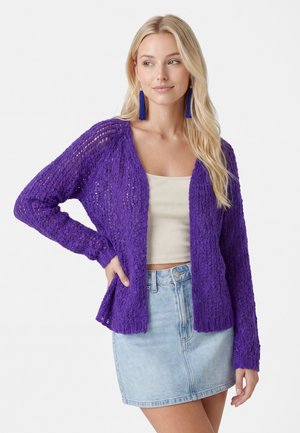 Lyshåret kvinde iført blå frynseørenringe, lilla åben strikket cardigan, beige crop top og lyse denim miniskørt, poserer med hånden på hoften.