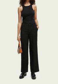 Zwarte wijde broek van soepel materiaal, met een knoopsluiting en zakken. Gecombineerd met een zwart geribbeld tanktop en een bruine tas.