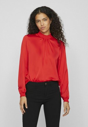 Red satin blouse with long sleeves, gathered neckline detail, and elasticized cuffs. Paired with black fitted pants. Smooth texture.