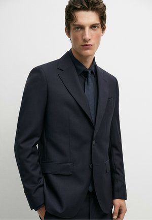 Jonge man met kort krullend haar, gekleed in een donker marineblauw pak, bijpassende stropdas en een zwart overhemd tegen een effen lichte achtergrond.