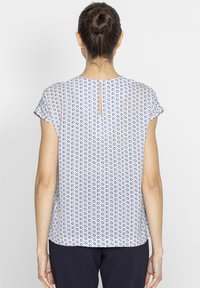 Bluzka z krótkim rękawem w kolorze białym z niebieskim geometrycznym wzorem. Posiada pionowe wycięcie w kształcie klucza z tyłu, miękką tkaninę oraz luźny krój.