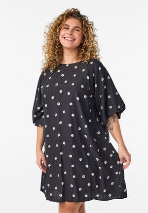 Usmievavá žena s kučeravými vlasmi v čiernych šatách po kolená s bielymi bodkami a voľnými trojštvrťovými rukávmi, ukazujúca kvetinový tetovanie na ruke.