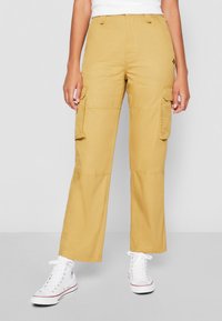 Osoba w żółtych spodniach cargo z kieszeniami na klapki i białych wysokich trampkach stoi na jednolitym tle.