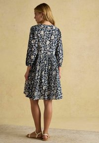 Eine junge Frau mit schulterlangen blonden Haaren trägt ein marineblaues Blumenkleid und beiges Sandalen, während sie vor einem strukturierten beigen Hintergrund steht.