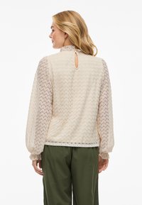 Krämfärgad spetsblus med långa, semi-transparenta ärmar och en nyckelhålsdetalj i ryggen, kombinerad med olivgröna byxor.